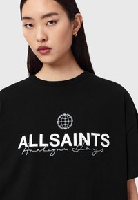 Νεαρή γυναίκα με μακριά σκούρα μαλλιά φοράει μαύρο T-shirt με λευκή επιγραφή "ALLSAINTS" και λογότυπο υδρόγειου πάνω από τη γραφική επιγραφή "Analogue Days".