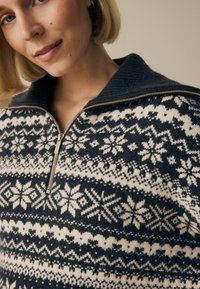 Gebreide trui met een marineblauwe basis en witte sneeuwvlok- en geometrische patronen. Een gouden rits en een donkere geribbelde kraag voegen detail toe.