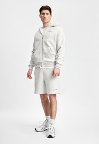 Lys grå hettejakke med glidelås i kombinasjon med matchende shorts, med lommer og subtil "GASPARA" merkevare i grønt. Brukt med hvite joggesko.