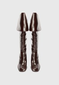 Par de botas de couro castanho escuro, até ao joelho, brilhantes, com largas golas dobradas e fechos laterais, apresentadas de frente sobre um fundo branco.