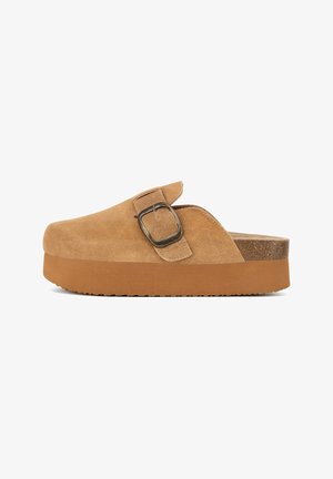 Tan suede clogs with a wide, rounded toe, a prominent metal buckle, and a thick, textured brown rubber sole for added grip.