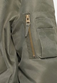 Olivenozeleni tkaninski odjevni predmet s kutićem na zip i zlatim detaljima te ravnim zakrpnim džepom, teksturirana površina.