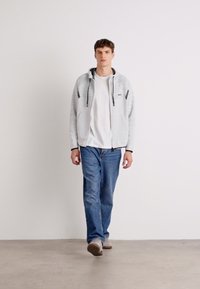 Hoodie zippé gris clair avec des accents noirs, porté sur un t-shirt blanc et associé à un jean bleu. Le modèle se tient devant un fond uni.