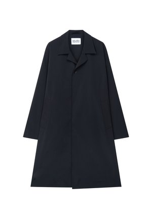 Junger Mann steht, trägt einen langen schwarzen Mantel, weißes Hemd, cremefarbene Hose und schwarze elegante Schuhe vor einem schlichten hellen Hintergrund.