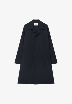 Jeune homme debout portant un long manteau noir, une chemise blanche, un pantalon couleur crème et des chaussures habillées noires sur un fond clair uni.