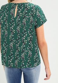 Blusa verde de manga corta con un patrón floral en tonos blanco y púrpura, detalle de apertura en la parte posterior y un corte relajado.
