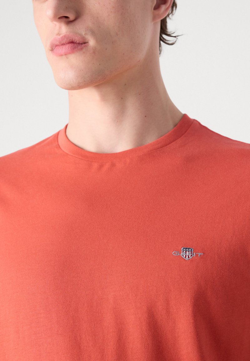 Kurzärmliges T-Shirt mit rundem Halsausschnitt aus korallfarbenem Stoff mit glatter Textur. Mit gesticktem Logo auf der linken Brust.