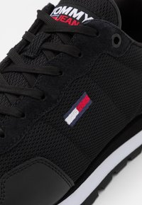 Baskets noires avec texture en mesh et daim, lacets noirs, semelle blanche, logo Tommy Jeans sur la languette et emblème de drapeau rouge, blanc et bleu sur le côté.