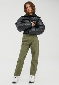 Giacca puffer in similpelle nera, pantaloni dritti verde oliva e sneakers bianche. Tessuto testurizzato, chiusura con zip, doppie tasche sul petto.