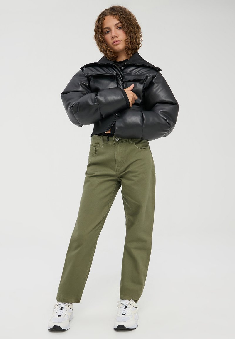 Giacca puffer in similpelle nera, pantaloni dritti verde oliva e sneakers bianche. Tessuto testurizzato, chiusura con zip, doppie tasche sul petto.