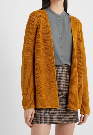 Femme portant un cardigan tricoté épais jaune moutarde sur une chemise grise à boutons et une mini-jupe marron à motif pied-de-poule.