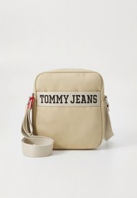 Bolso bandolera cuadrado beige con la inscripción "TOMMY JEANS" impresa en el centro frontal y una correa de hombro beige ajustable.