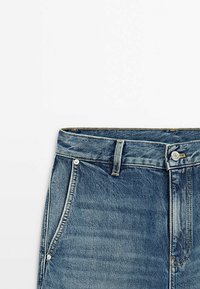 Jeans in denim con lavaggio blu, caratterizzati da un design a cinque tasche, passanti per cintura e chiusura con bottone in metallo in vita.