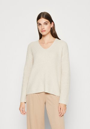 Beżowy sweter dzianinowy z dekoltem w serek, textured wzorem, szerokimi rękawami i luźnym krojem. Połączony z jasnobeżowymi spodniami.