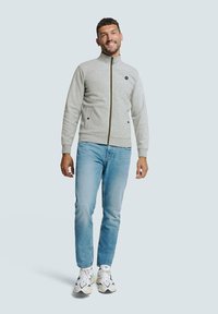 Grijze zip-up jas met een gestructureerd patroon, twee voorzakken en donkere accenten, gedragen met lichtblauwe jeans en witte sneakers.