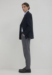 Blazer blu navy con taglio su misura, pantaloni grigi e scarpe in pelle nera con doppia fibbia. Camicia azzurra sotto.