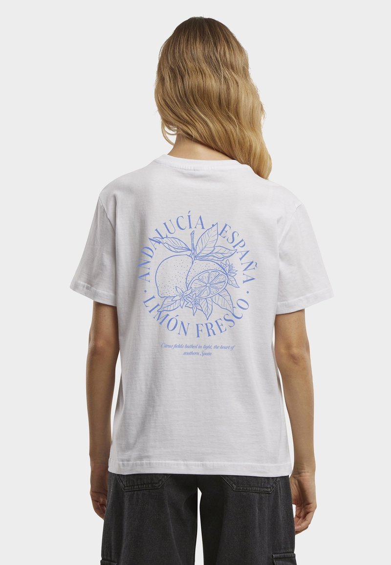Frau mit langen blonden Haaren trägt ein weißes T-Shirt mit blauer Zitronenillustration und dem Text „Andalucía España Limón Fresco“ auf der Rückseite.
