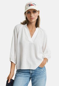 Witte linnen blouse met een V-hals en driekwart mouwen, met gathered manchetten. Gecombineerd met lichtblauwe spijkerbroeken en een crèmekleurige pet met geborduurde tekst.