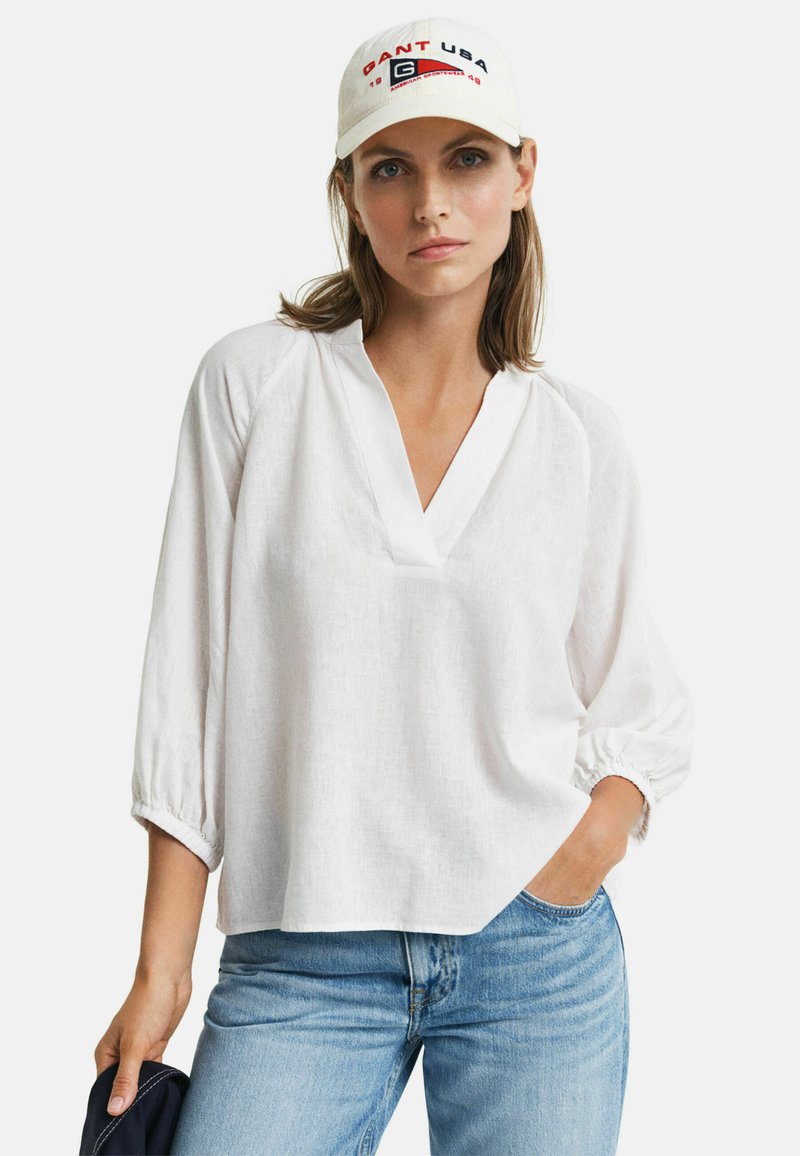 Witte linnen blouse met een V-hals en driekwart mouwen, met gathered manchetten. Gecombineerd met lichtblauwe spijkerbroeken en een crèmekleurige pet met geborduurde tekst.