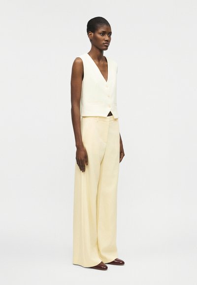 Γυναίκα που στέκεται φορώντας αμάνικο λευκό γιλέκο με κουμπιά και κρεμ παντελόνια φαρδιάς γραμμής με καφέ παπούτσια, πάνω σε απλό φωτεινό φόντο.