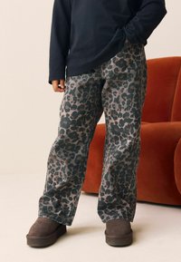 Løstsiddende leopardprint bukser i mørkegrønne og brune toner, lavet af et tekstureret stof. Båret med en langærmet sort top og mørke støvler.