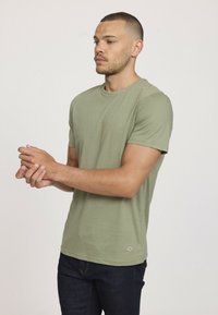 T-shirt à manches courtes olive en tissu doux avec un col rond ; présente un petit logo près de l'ourlet et est associé à un jeans foncé.
