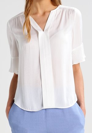 Hvid V-hals bluse med trekvartlange ærmer og frontplisseringsdetalje, båret stoppet ned i lyseblå bukser.