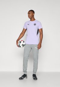 Camisa corta de manga corta morada clara con detalles y logotipo en negro, pantalones de chándal grises y zapatillas deportivas negras, sosteniendo un balón de fútbol en blanco y negro.