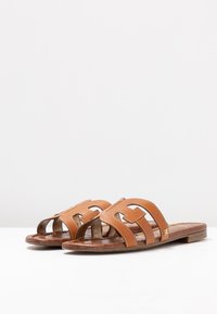 Sandalias de cuero marrón con correas entrelazadas, diseño de punta abierta y suela plana. Plantilla texturizada con un patrón, detalle sutil de hardware en el lateral.