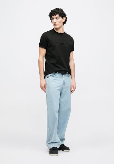 Μαύρο βαμβακερό μπλουζάκι με ανάγλυφο λογότυπο, σε συνδυασμό με ανοιχτό μπλε παντελόνι με φαρδιά μπατζάκια. Μαύρα αθλητικά παπούτσια με υφασμένες σόλες ολοκληρώνουν το σύνολο.