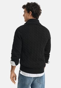 Black cable-knit sweater with a high collar, ribbed cuffs, and hem, paired with a striped shirt underneath. Visible denim jeans.