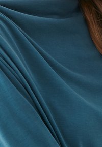 Turkoosi kangas laskeutuu pehmeästi sileän tekstuurin kanssa. Materiaali näyttää kevyeltä ja hieman joustavalta, ja siinä on hienovaraisia taitoksia ja ryppyjä.