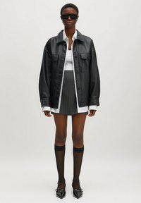 Schwarze Leder-oversized-Jacke mit grauem Kragen, darunter ein weißes Hemd, grauer Nadelstreifen-Minirock, durchsichtige schwarze knielange Socken, spitze schwarze Absätze.