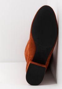 Botín de ante en naranja quemado con suela de goma negra. Presenta una puntera redonda y tacón cuadrado, con líneas limpias y acentos mínimos.