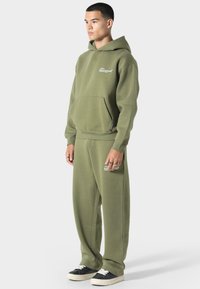 Oliivinvihreä huppari ja sen mukaiset verkkarit pehmeästä kankaasta, joissa on etutasku ja logoyksityiskohta. Mustat tennarit täydentävät asukokonaisuuden.