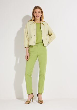 Frau steht und trägt ein hellgrünes Oberteil, passende grüne Hose, beige kurze Jacke und olivgrüne Sandalen mit hohen Absätzen, vor weißem Hintergrund.