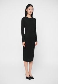 Γυναίκα που στέκεται φορώντας ένα εφαρμοστό, μακρυμάνικο μαύρο μίντι φόρεμα και μαύρες μυτερές γόβες σε απλό λευκό φόντο.