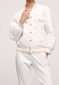 Quilted white bomber jacket with gold buttons, features a chest pocket and ribbed cuffs. Worn over matching white pants with gold accents.