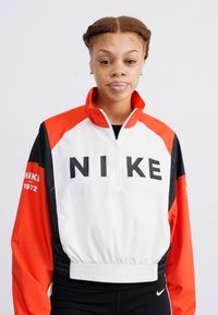 Balts un sarkans Nike puloverjakas ar pusē iestrādātu rāvējslēdzēju, melnu logotipa tekstu, elastīgu jostu un oranžām piedurknēm. Valkā melnas sporta legingas.