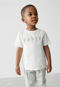 Valkoinen puuvillainen t-paita, jossa on harmaalla tekstillä "BAKER", yhdistetty vaaleanharmaisiin mallikuviollisiin housuihin. Sileä pinta, pyöreäkauluksinen malli.