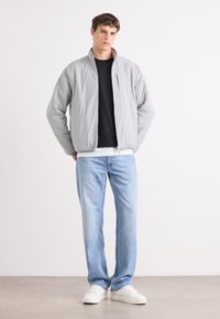 Grå polstret jakke med ståkrage, svart genser under, lyseblå jeans og hvite sneakers på en nøytral bakgrunn.