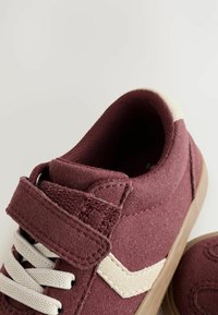 Maroonsko med velcro-strop og beige accenter, med snørebånd og en tekstureret overflade, placeret mod en lys baggrund.