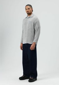 Grijze pluche lange mouwen polo met kraag en geborduurd blauw logo, gecombineerd met een donkere spijkerbroek en zwarte schoenen met dikke zolen.