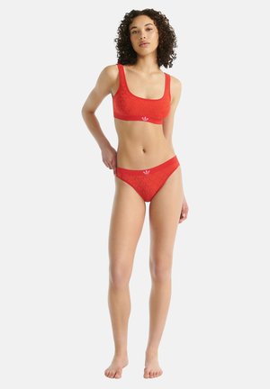 Mujer con cabello rizado de pie descalza, vistiendo un sujetador deportivo rojo de Adidas y bragas a juego con el logo sobre un fondo liso.