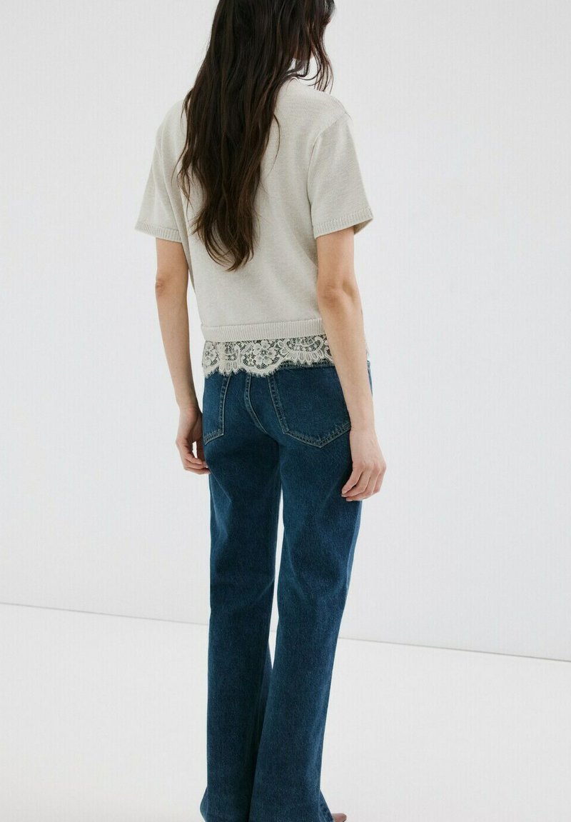 Mujer con cabello largo y oscuro, vestida con una blusa beige de manga corta con borde de encaje y jeans azul oscuro, de pie frente a un fondo blanco.