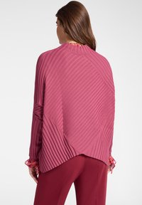 Mujer vestida con un suéter asimétrico de canalé rosa con mangas largas y pantalones burdeos, vista desde atrás sobre un fondo liso.