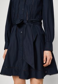 Abito camicia blu navy con maniche lunghe, chiusura a bottoni frontale, vita strutturata con cintura e dettagli in pizzo sull'orlo. Tessuto morbido e leggero.