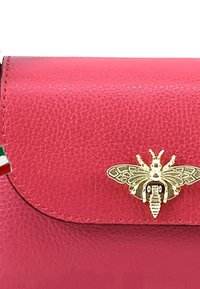 Sac en cuir rouge à texture granuleuse avec un emblème doré en forme d'abeille sur le rabat, présentant de fines coutures et un petit charm de drapeau italien accroché.