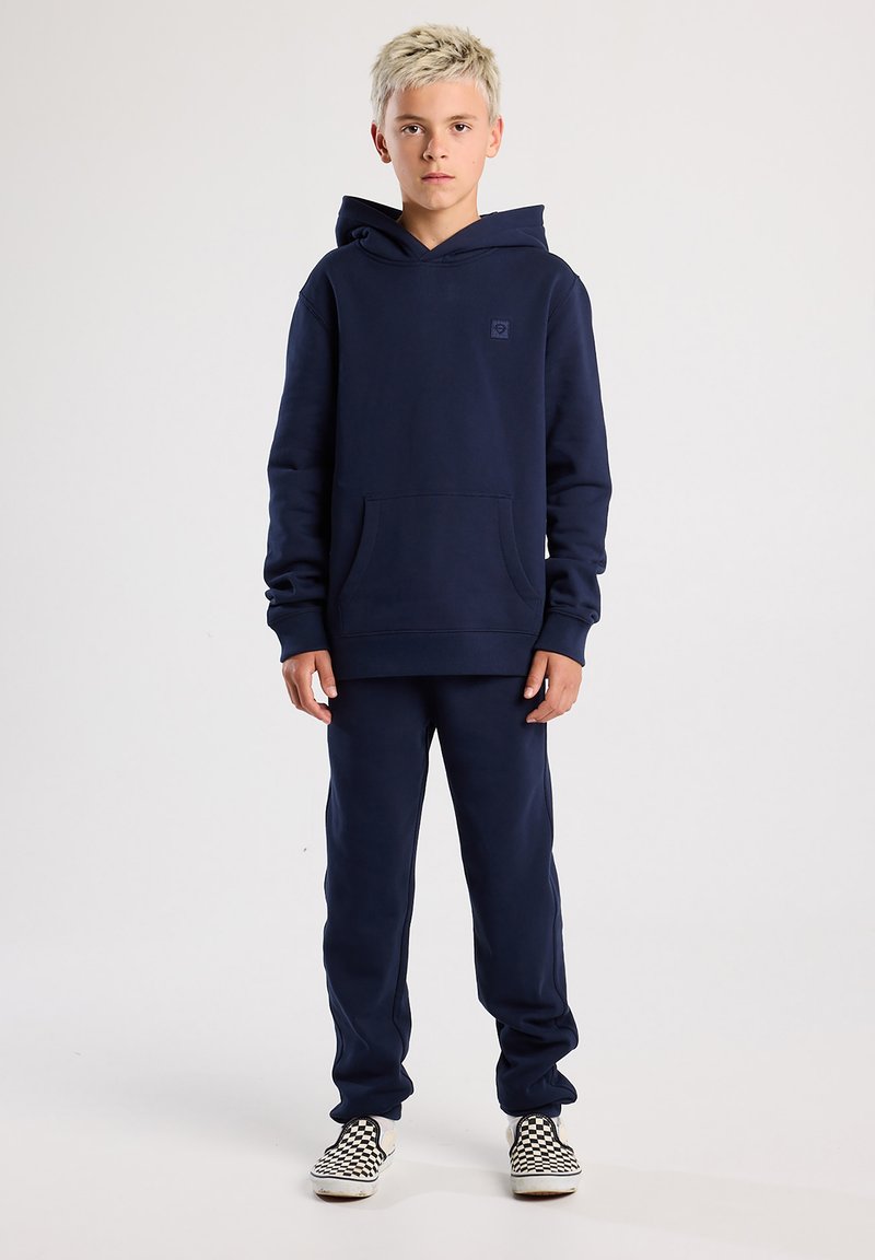Blond jongetje dat naar voren kijkt, draagt een marineblauwe hoodie, bijpassende joggingbroek en zwart-witte geruite slip-on schoenen op een effen achtergrond.