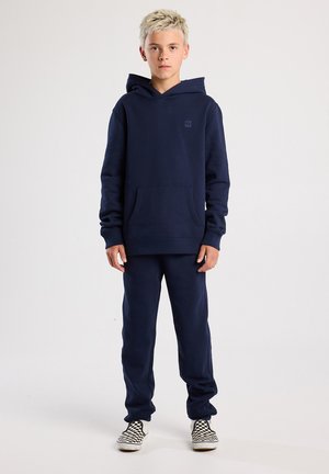Sudadera con capucha de color azul marino y pantalones deportivos a juego, hechos de una tela suave. Cuenta con un bolsillo delantero y puños elásticos; se lleva con zapatos a cuadros en blanco y negro.
